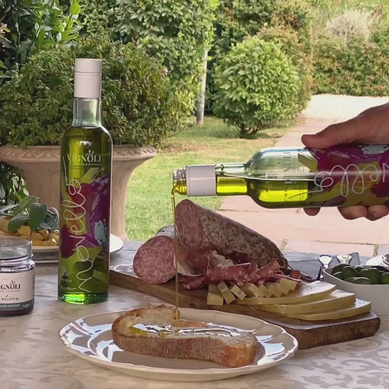 Vignoli Novello Extra Virgin Olive Oil front of 16.9oz bottle being poured over bread with charcuterie behind