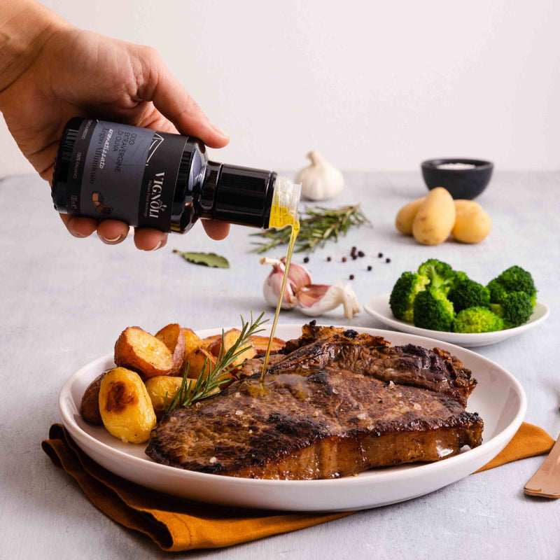 Vignoli Mesquite Smoked Infused Extra Virgin Olive Oil front of 8.5 oz bottle pouring over steak and potatoes with broccoli in the background